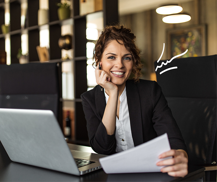 junge Frau im schwarzen Business-Outfit sitzt vor dem Laptop und hält ein Papier in der Hand